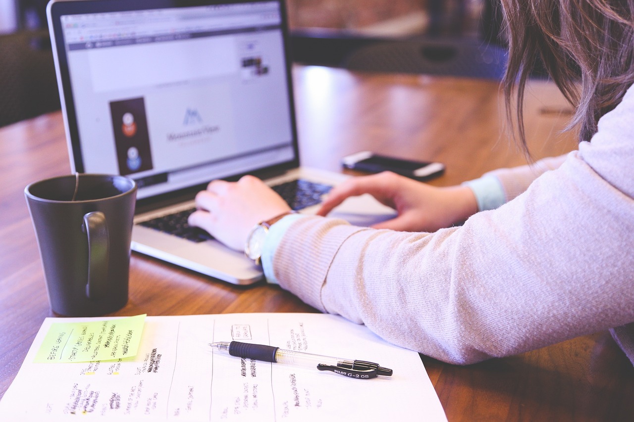 Woman typing on a laptop with papers and a pen sitting beside her on a desk.